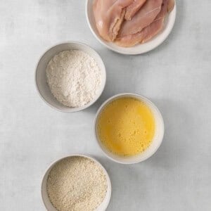 four bowls with chicken, flour, eggs and oil on a grey background.