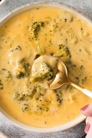 a bowl of broccoli cheese soup with a hand holding a spoon.