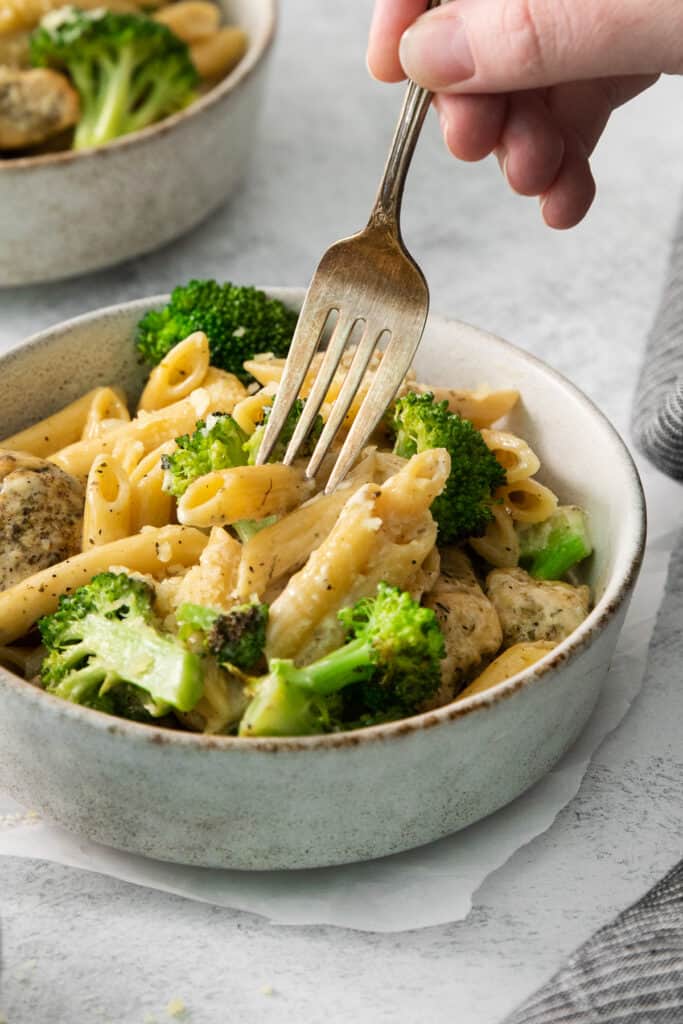 a person holding a fork while eating a bowl of pasta with chicken and broccoli.