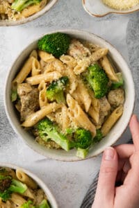 a person holding a bowl of pasta with chicken and broccoli.