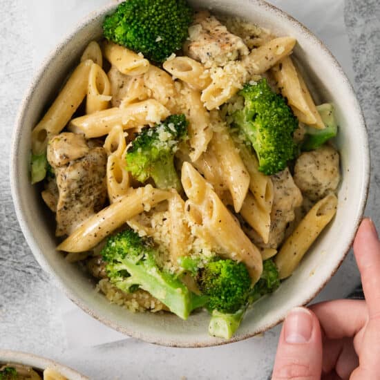 a person holding a bowl of pasta with chicken and broccoli.