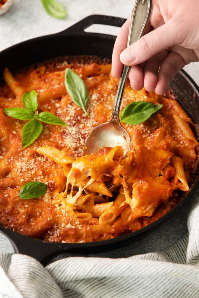 a hand holding a spoon over a skillet of pasta.
