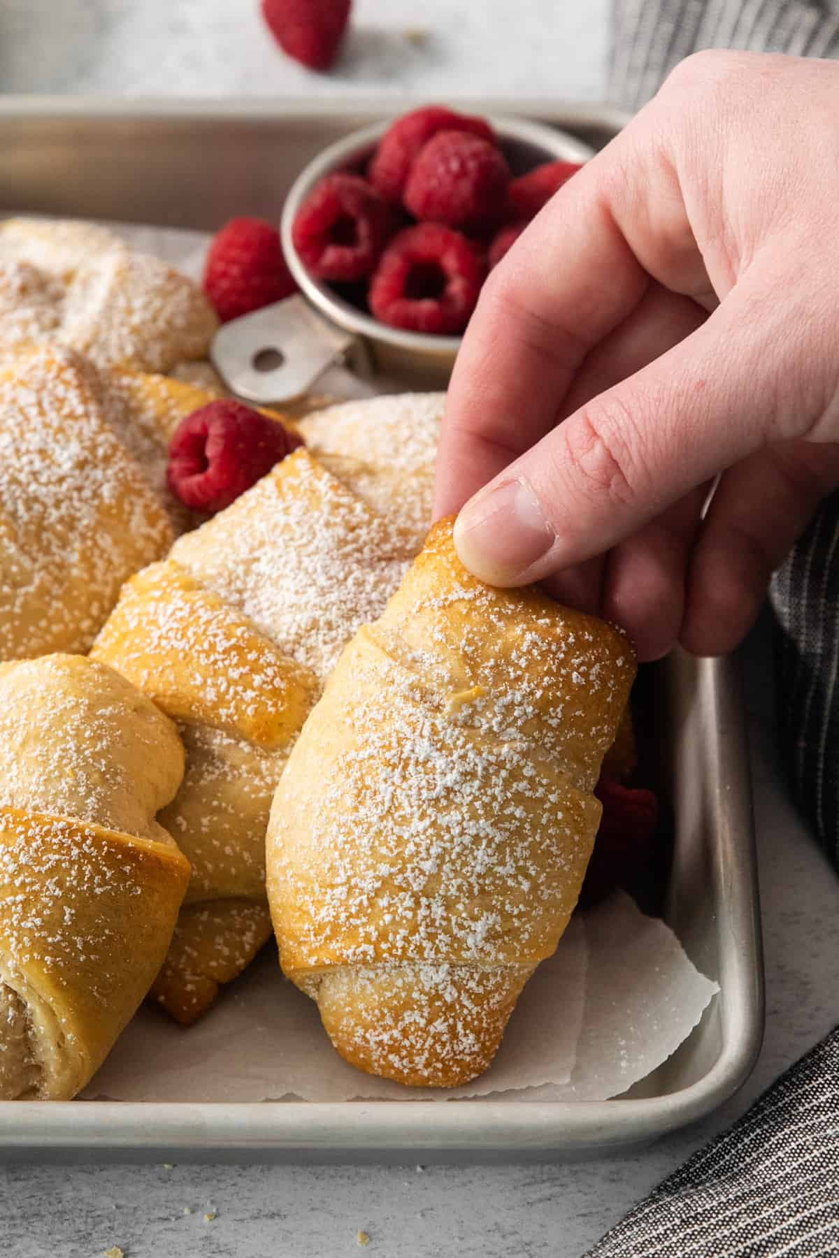 a hand grabbing a cinnamon roll with raspberries on a baking sheet.