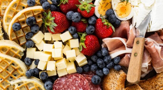 A platter with a variety of meats, cheeses, fruits and waffles.