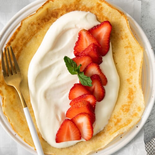 A crepe topped with a luscious cream cheese crepe filling, sliced strawberries, and a sprig of mint, served on a white plate with a gold and white fork.