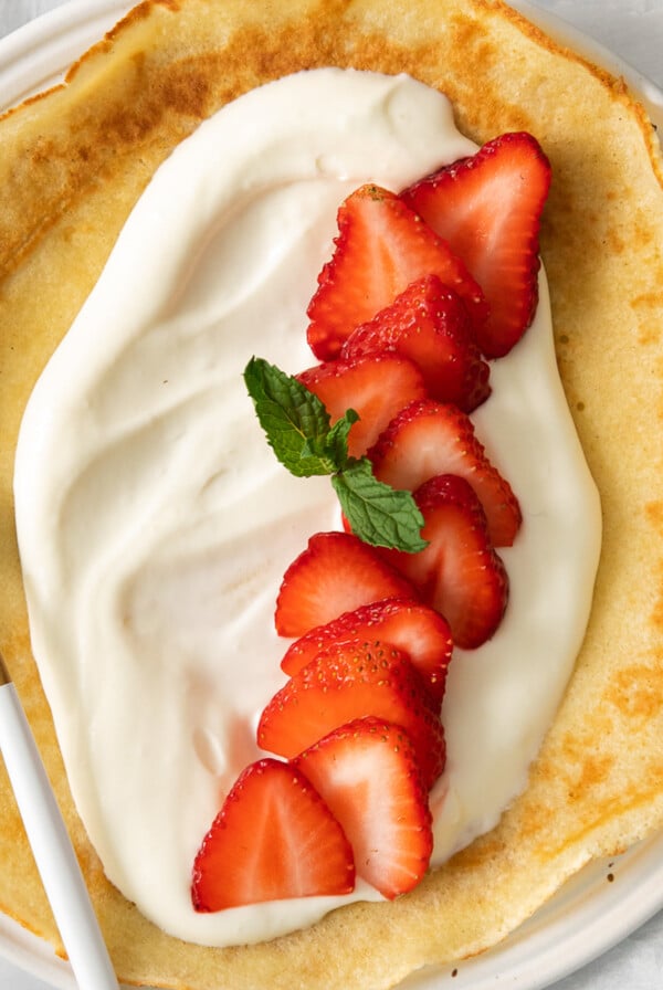 A crepe topped with a luscious cream cheese crepe filling, sliced strawberries, and a sprig of mint, served on a white plate with a gold and white fork.