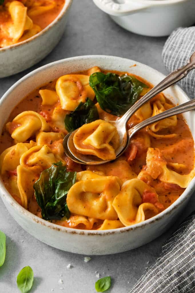 A bowl of creamy tomato tortellini soup garnished with spinach, served with a spoon.