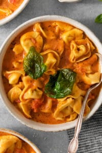 A bowl of tomato-based tortellini soup garnished with basil leaves.