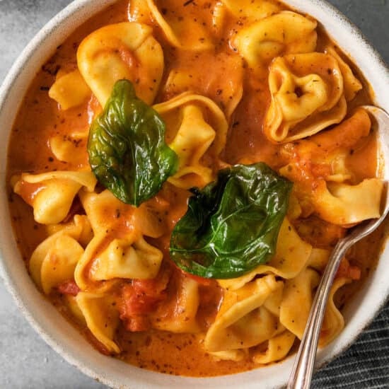 A bowl of tomato-based tortellini soup garnished with basil leaves.