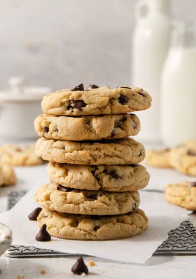 A stack of five egg-free chocolate chip cookies is placed on a napkin, with more cookies and bottles of milk in the blurred background.