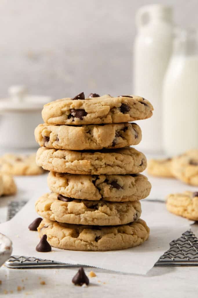 A stack of five egg-free chocolate chip cookies is placed on a napkin, with more cookies and bottles of milk in the blurred background.