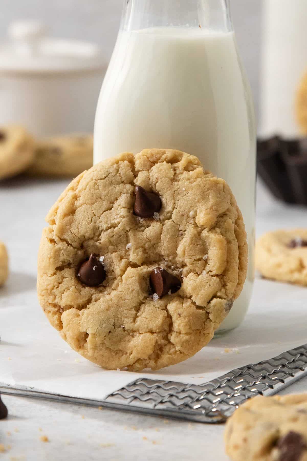 An egg-free chocolate chip cookie is propped against a glass bottle of milk. Several more cookies and baking racks are in the background.