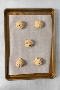 A baking sheet lined with parchment paper holds five scoops of egg-free chocolate chip cookie dough evenly spaced and ready for baking.