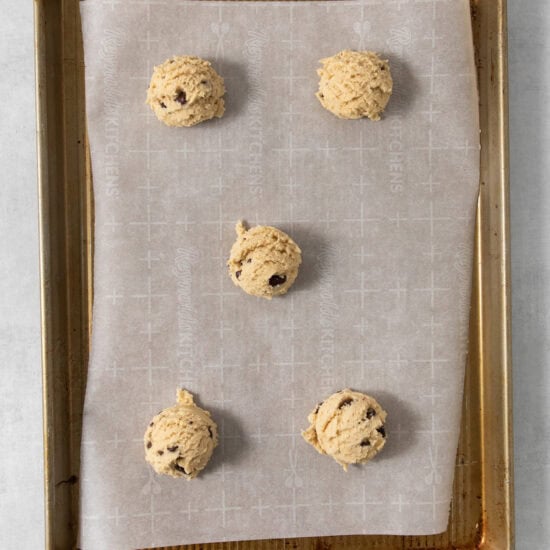 A baking sheet lined with parchment paper holds five scoops of egg-free chocolate chip cookie dough evenly spaced and ready for baking.