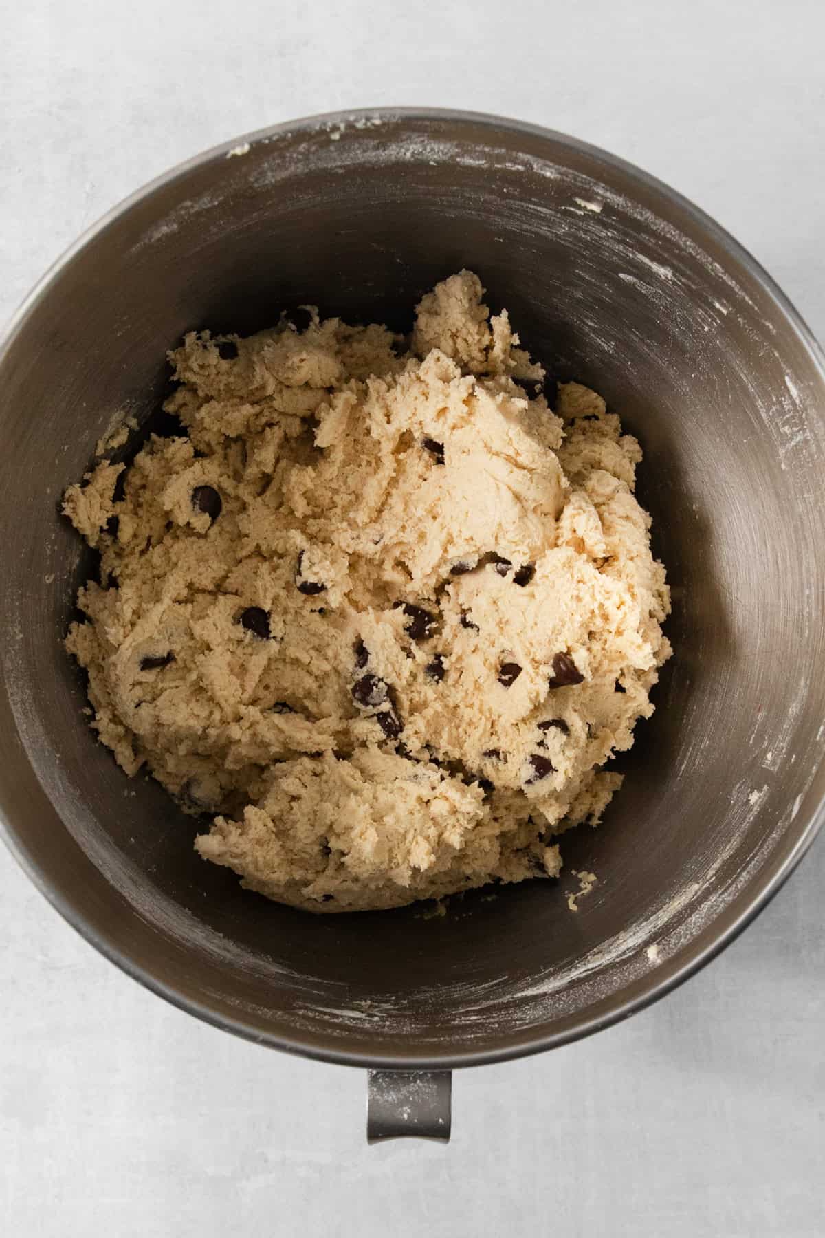 A metal mixing bowl brimming with egg-free chocolate chip cookie dough rests on a light gray background.