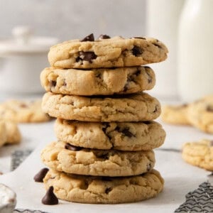 A stack of five egg-free chocolate chip cookies on a white surface, with more cookies and blurred bottles in the background.