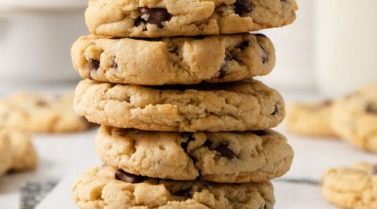 A stack of five egg-free chocolate chip cookies on a white surface, with more cookies and blurred bottles in the background.
