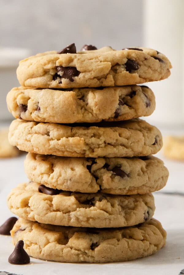 A stack of five egg-free chocolate chip cookies on a white surface, with more cookies and blurred bottles in the background.