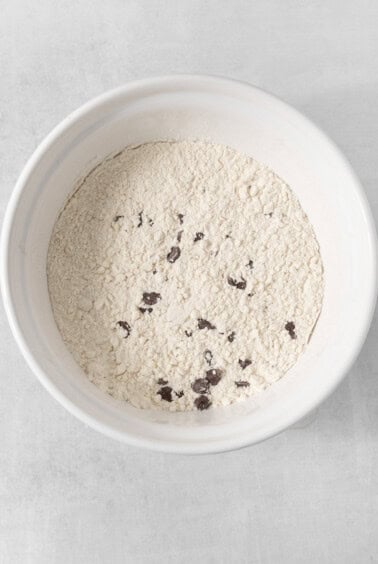 A white bowl is filled with a mixture of light-colored flour and small, dark chunks, likely chocolate. This blend suggests the beginnings of egg-free chocolate chip cookies. The surface is unevenly distributed and the bowl rests on a light-colored surface.