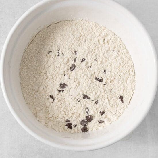 A white bowl is filled with a mixture of light-colored flour and small, dark chunks, likely chocolate. This blend suggests the beginnings of egg-free chocolate chip cookies. The surface is unevenly distributed and the bowl rests on a light-colored surface.