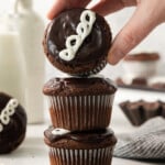 A hand is holding a chocolate cupcake with white icing, stacking it on two other similar cupcakes. The background includes a milk bottle, grey cloth, and a spoon.