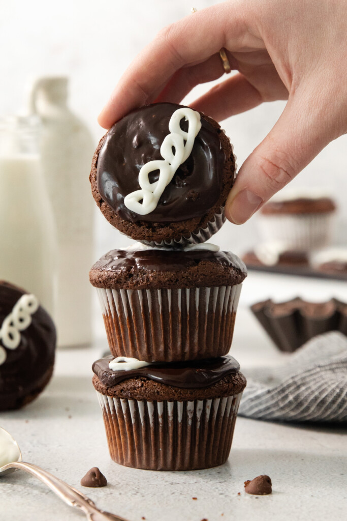 A hand is holding a chocolate cupcake with white icing, stacking it on two other similar cupcakes. The background includes a milk bottle, grey cloth, and a spoon.