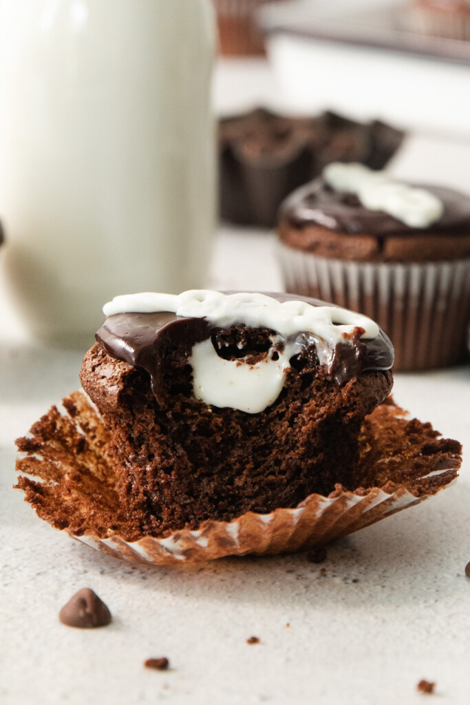 A chocolate cupcake with a bite taken out, revealing a white cream filling, sits on a countertop with its wrapper partially open. A milk bottle and more cupcakes are in the background.