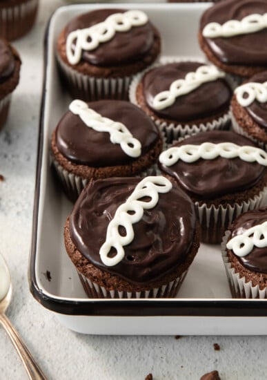 A tray filled with chocolate cupcakes topped with a swirl of white icing, neatly arranged in rows. A spoon and additional cupcakes are partially visible in the background.