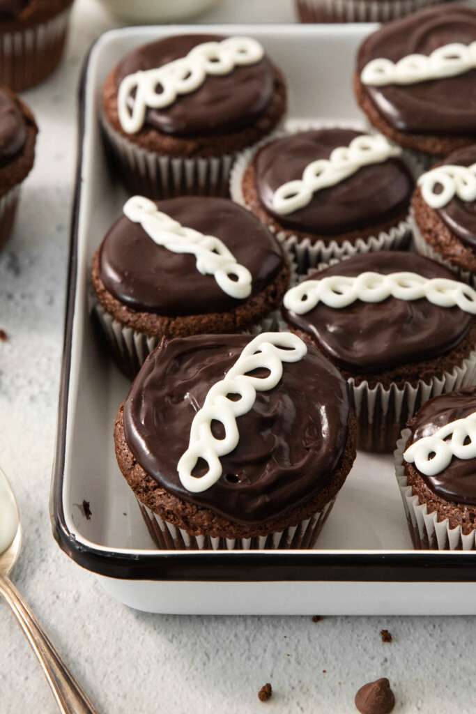 A tray filled with chocolate cupcakes topped with a swirl of white icing, neatly arranged in rows. A spoon and additional cupcakes are partially visible in the background.