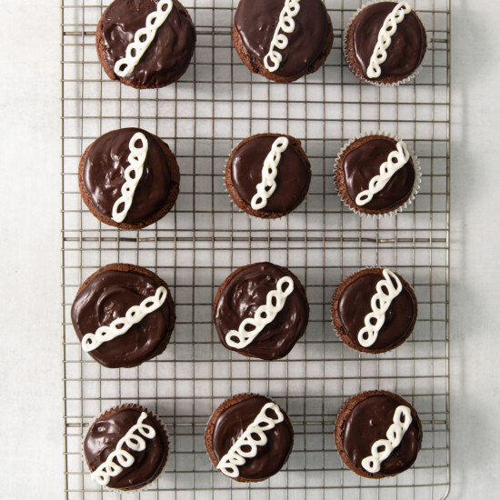 A cooling rack with twelve chocolate-covered cupcakes, each topped with white icing squiggles, arranged in three rows of four.