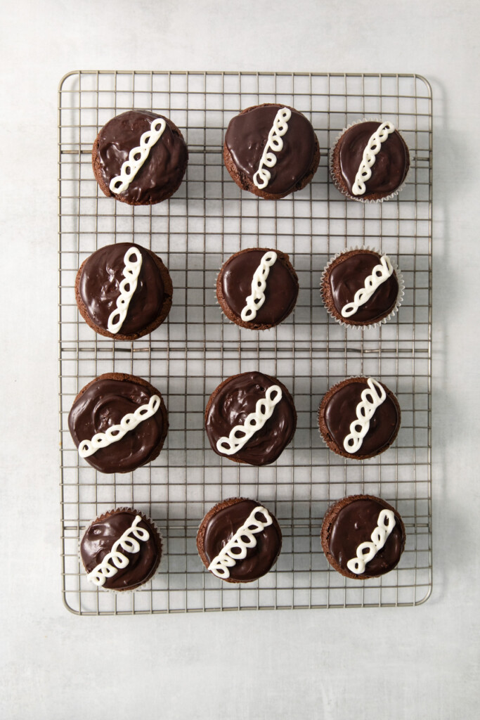 A cooling rack with twelve chocolate-covered cupcakes, each topped with white icing squiggles, arranged in three rows of four.