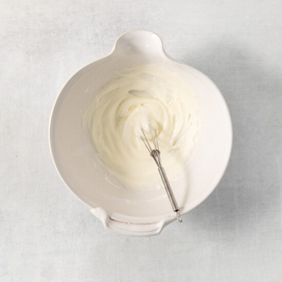 A small metal whisk sits in a white bowl containing a creamy mixture on a light grey surface.