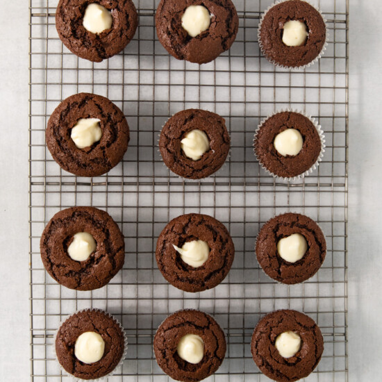 A cooling rack with twelve chocolate muffins, each with a dollop of white filling in the center.