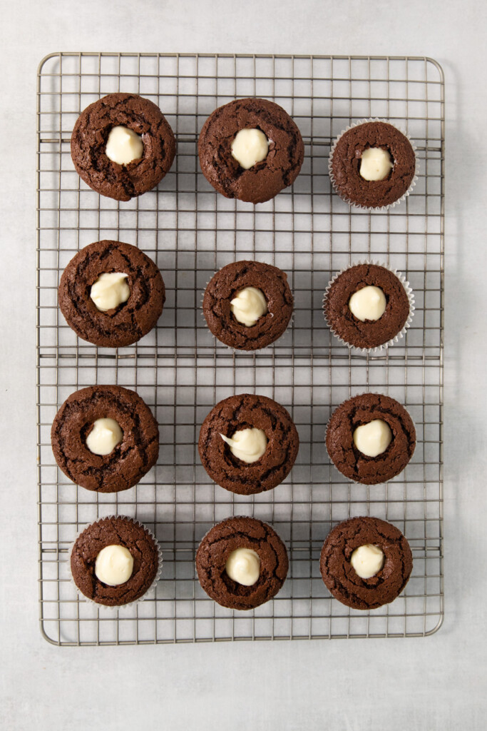 A cooling rack with twelve chocolate muffins, each with a dollop of white filling in the center.