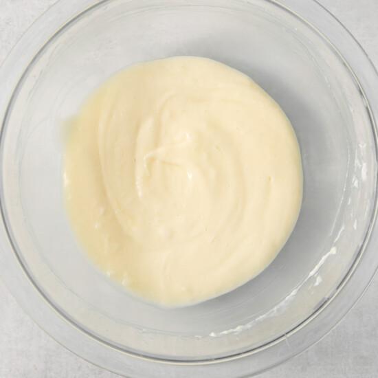 A glass bowl containing a smooth, creamy mixture, likely batter or dough, is positioned against a light gray background.