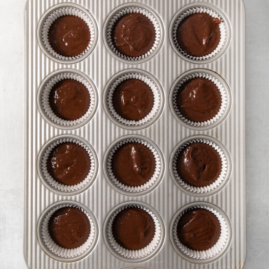 A muffin tin filled with twelve paper liners containing chocolate batter, ready for baking.