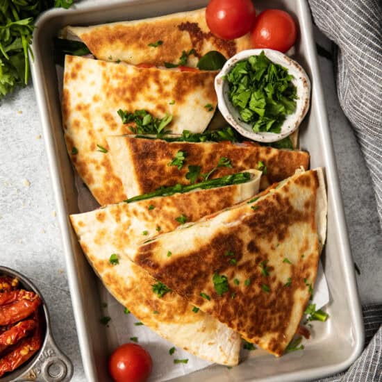 A tray of folded, golden-brown quesadillas garnished with chopped cilantro and accompanied by cherry tomatoes and a small bowl of chopped greens.