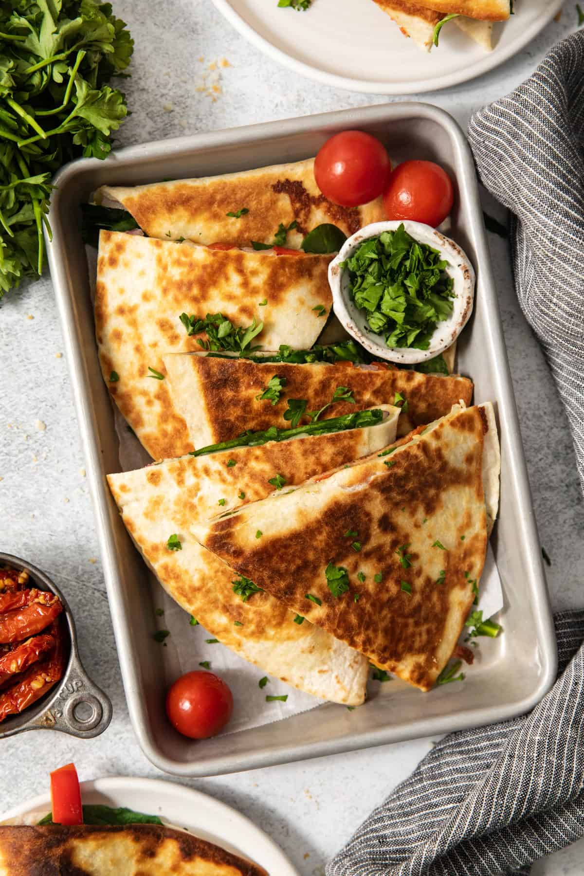 A tray of folded, golden-brown quesadillas garnished with chopped cilantro and accompanied by cherry tomatoes and a small bowl of chopped greens.