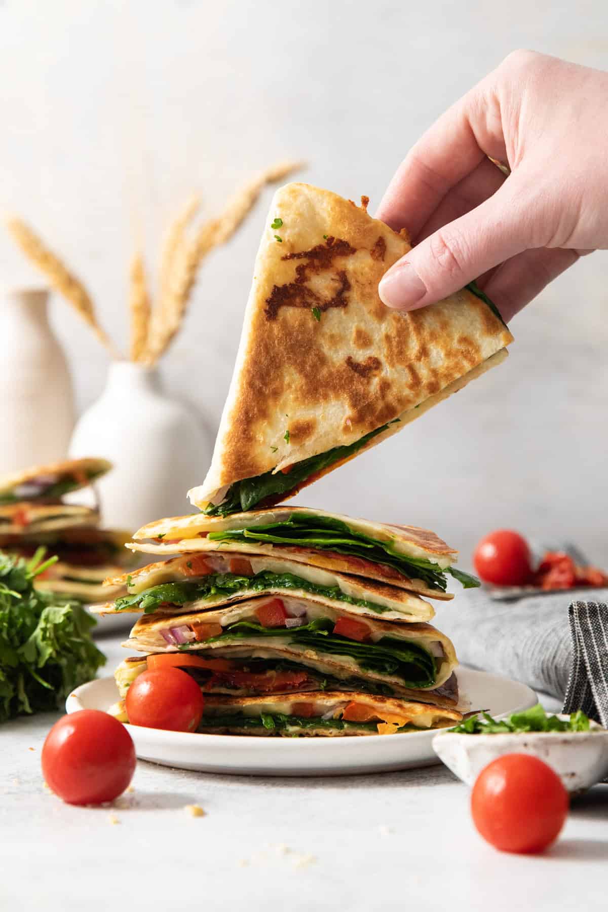 A hand lifting a slice from a stack of quesadillas filled with vegetables, with tomatoes, greens, and decorative items in the background.