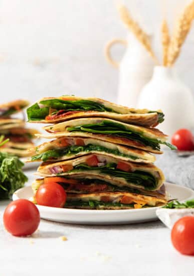 A stack of sliced vegetable quesadillas on a white plate, surrounded by cherry tomatoes and fresh spinach, with a blurred background of vases and wheat stalks.