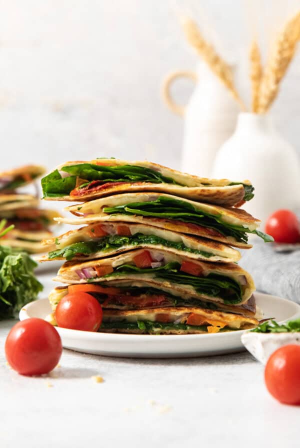 A stack of sliced vegetable quesadillas on a white plate, surrounded by cherry tomatoes and fresh spinach, with a blurred background of vases and wheat stalks.