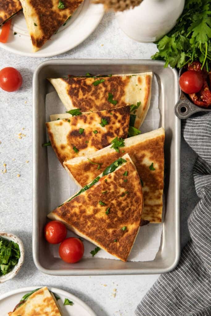 A metal tray with browned quesadilla slices garnished with chopped herbs, accompanied by whole cherry tomatoes. Fresh greens and tomatoes are visible nearby.