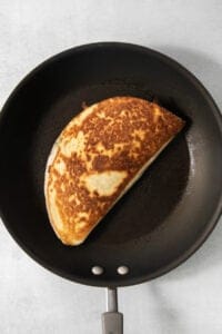A browned, folded quesadilla is cooking in a black non-stick frying pan.