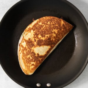 A browned, folded quesadilla is cooking in a black non-stick frying pan.