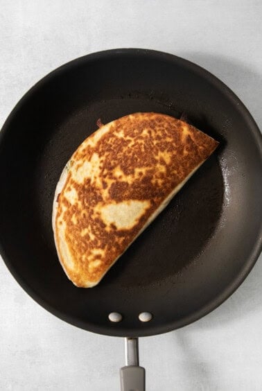 A browned, folded quesadilla is cooking in a black non-stick frying pan.