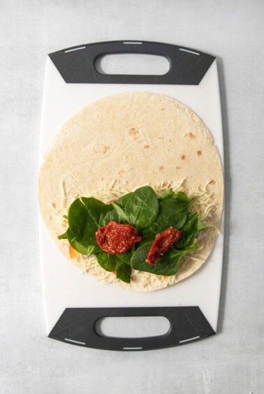 A tortilla with shredded cheese, leafy greens, and sun-dried tomatoes placed on a cutting board.