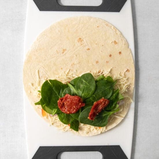 A tortilla with shredded cheese, leafy greens, and sun-dried tomatoes placed on a cutting board.