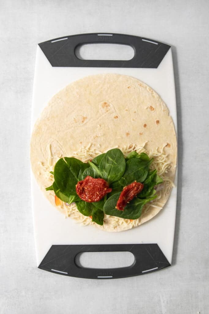 A tortilla with shredded cheese, leafy greens, and sun-dried tomatoes placed on a cutting board.