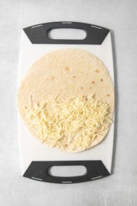 A tortilla partially filled with shredded cheese is placed on a white and black cutting board.