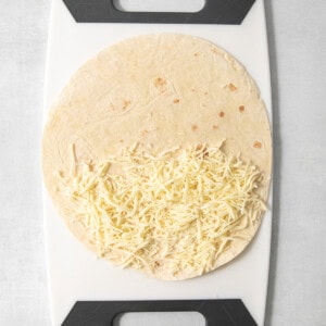 A tortilla partially filled with shredded cheese is placed on a white and black cutting board.